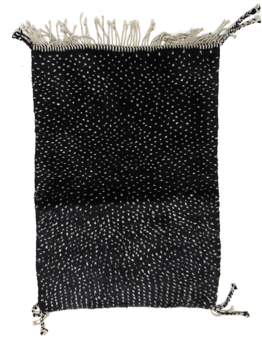 Hand-loomed black wool area rug featuring a speckled white-and-cream confetti texture, with a chunky top fringe and braided corner tassel ties, displayed on a white background.

