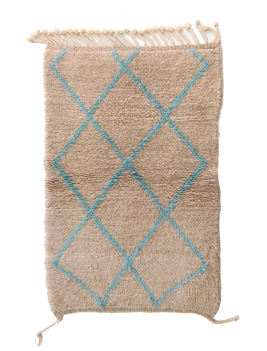 Plush handwoven wool rug in warm sand beige featuring a large overlapping diamond lattice pattern in muted teal/aqua, with a knotted fringe at the top and small corner ties, displayed on a white background