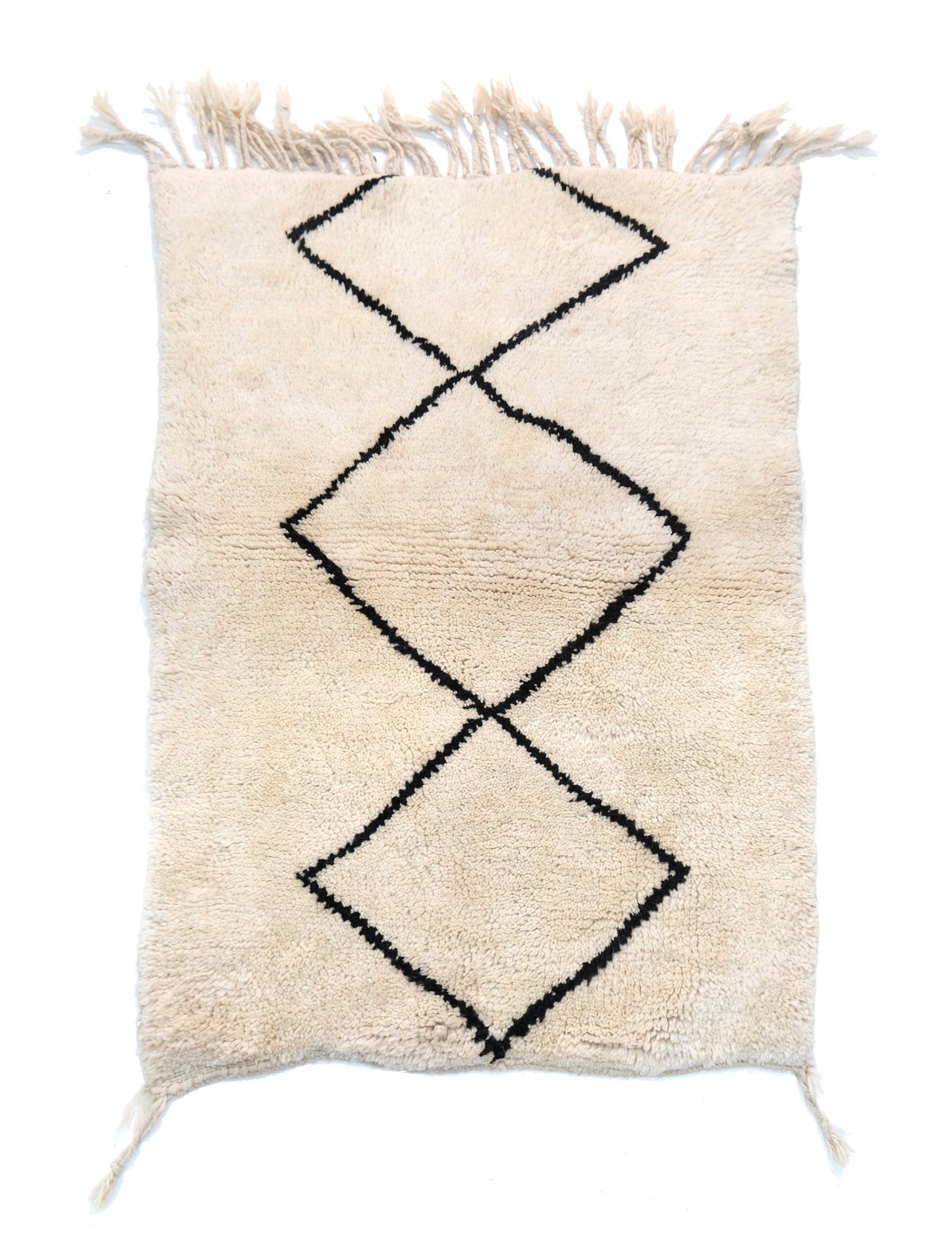 A cream-colored handwoven Moroccan shag rug featuring a large, central interlocking black diamond motif, with cream fringe detailing at the top and bottom edges and corner accents.

