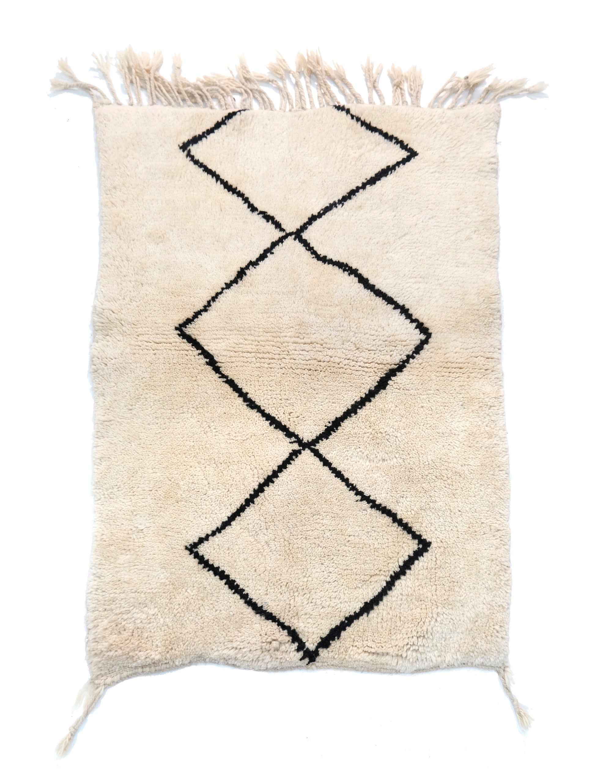A cream-colored handwoven Moroccan shag rug featuring a large, central interlocking black diamond motif, with cream fringe detailing at the top and bottom edges and corner accents.

