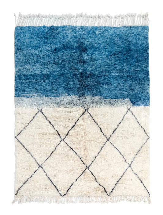 A handwoven Moroccan shag rug featuring a blue color-block top section blending into a cream bottom section with a black diamond trellis pattern, finished with white fringe on the top and bottom edges.

